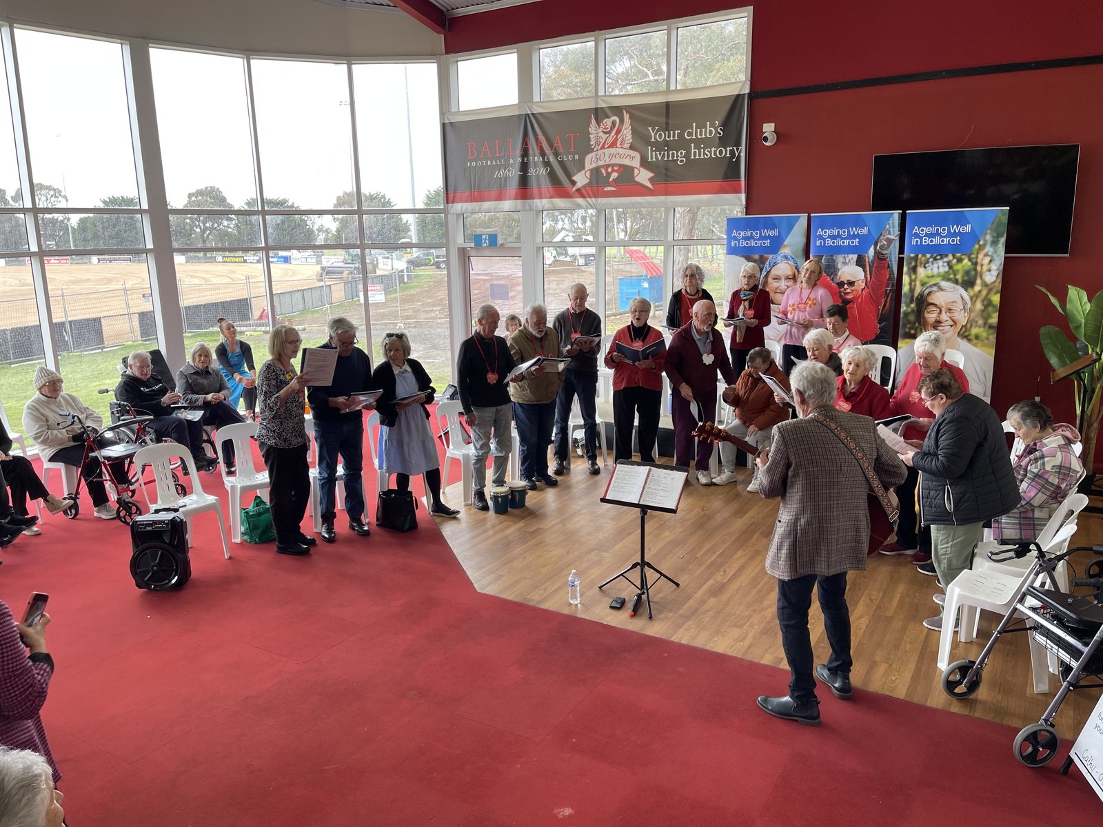 The Bigger Hearts Choir performing at the Ballarat Football Club. Members stand together with songbooks while musician Geoff Hassall leads on guitar.