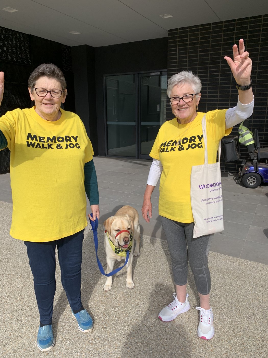 Two women in bright yellow Memory Walk and Jog t-shirts, arms raised in celebration, with their dementia assistance dog wearing a matching yellow bandana.